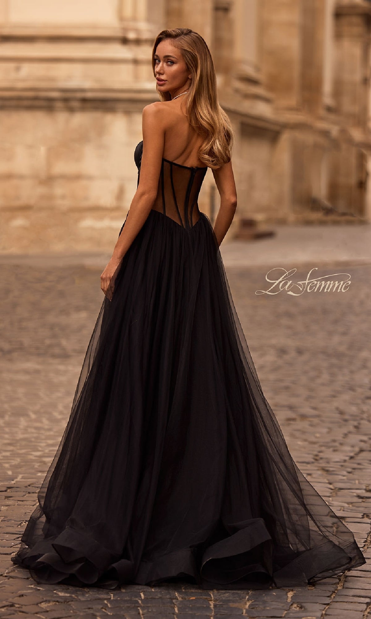 Wearing the La Femme 32771, a black strapless long prom dress with a sheer corset back, a woman stands on a cobblestone street and looks over her shoulder.